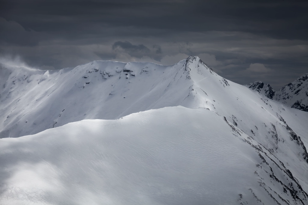 Cimes des Pyrénées enneigées où les rayons de soleil lancent un dernier assaut de lumière pour survivre avant la tempête, photographie d'art de Jill Pollock, Vaea Designs
