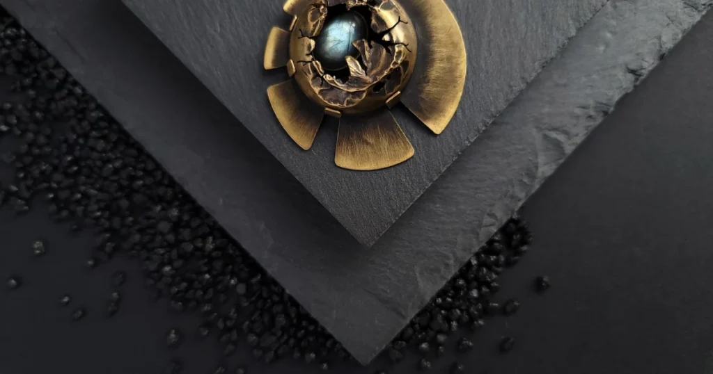 Broche nordique en laiton et labradorite posée sur fond ardoise et sable volcanique. Photo de couverture du livre "L'Art de la photographie de bijoux artisanaux" qui enseigne comment photographier ses bijoux.