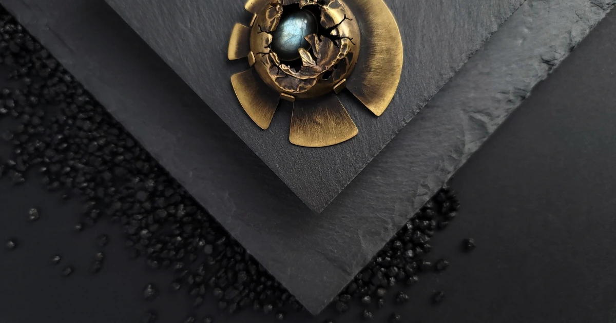 Broche nordique en laiton et labradorite posée sur fond ardoise et sable volcanique. Photo de couverture du livre "L'Art de la photographie de bijoux artisanaux" qui enseigne comment photographier ses bijoux.